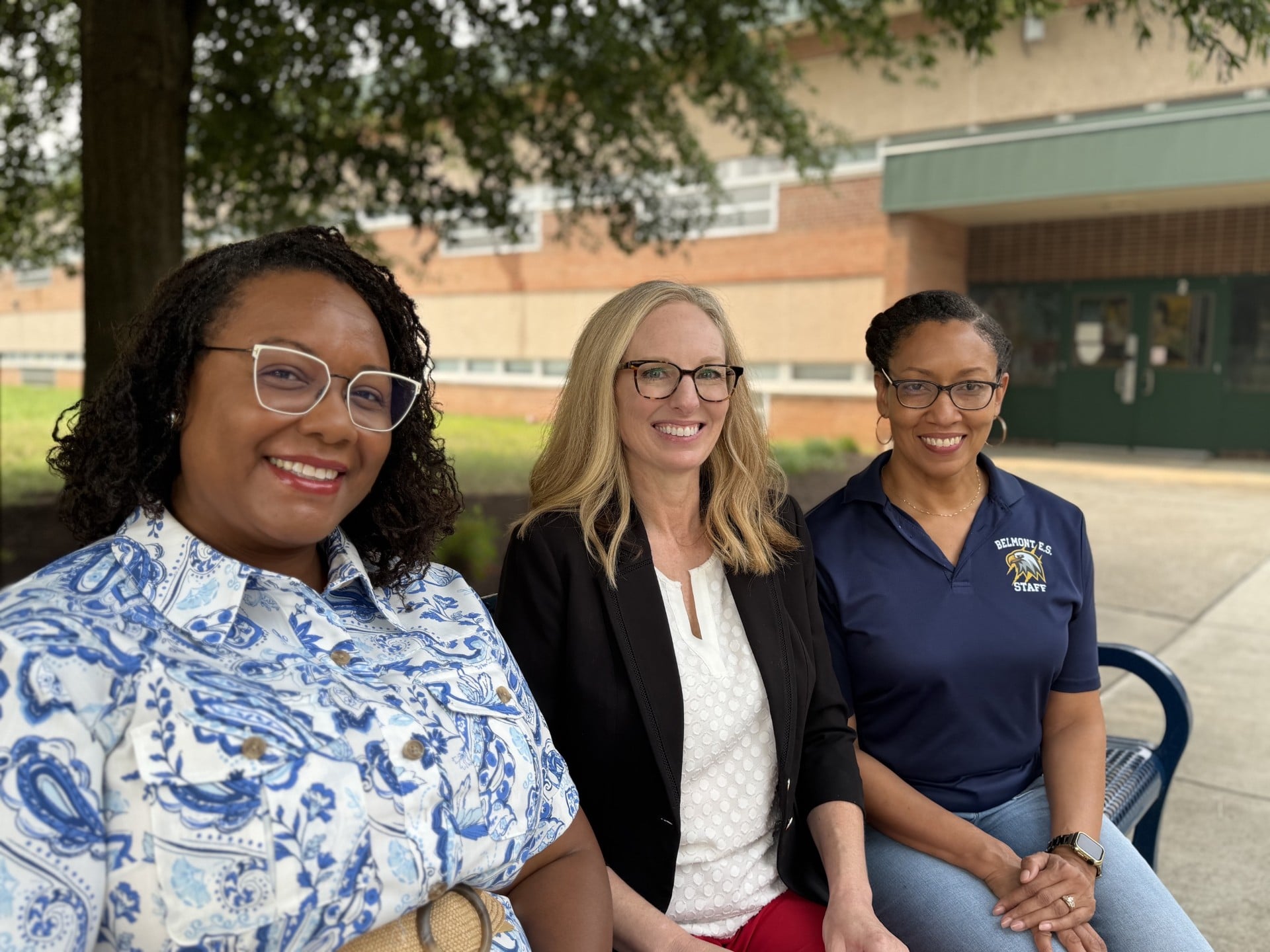 Three women smiling while seated outdoors in front of a school building; one wears a Belmont E.S. staff polo.